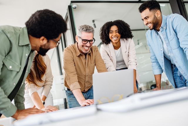 A cheerful team of diverse coworkers lean in around a laptop working on a project