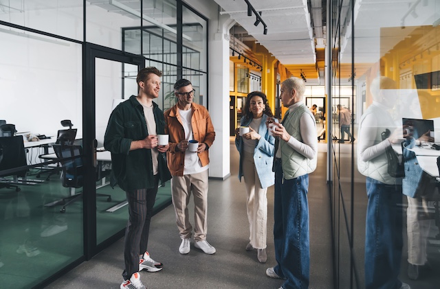 A group of diverse colleagues talk in a hallway as they hold their coffee mugs