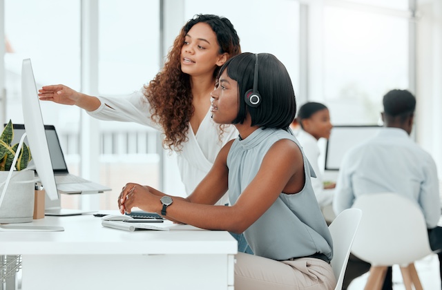 A young woman helps her female colleague at her desk